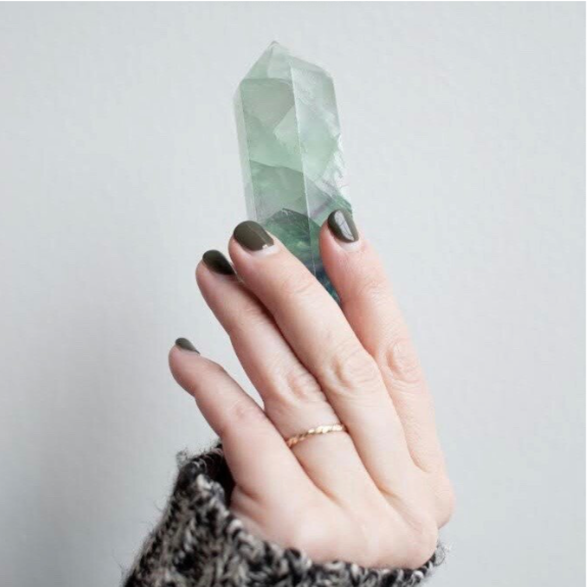 A hand wearing a slim gold Light + Grace band, holding a pale green fluorite crystal against a soft grey background.